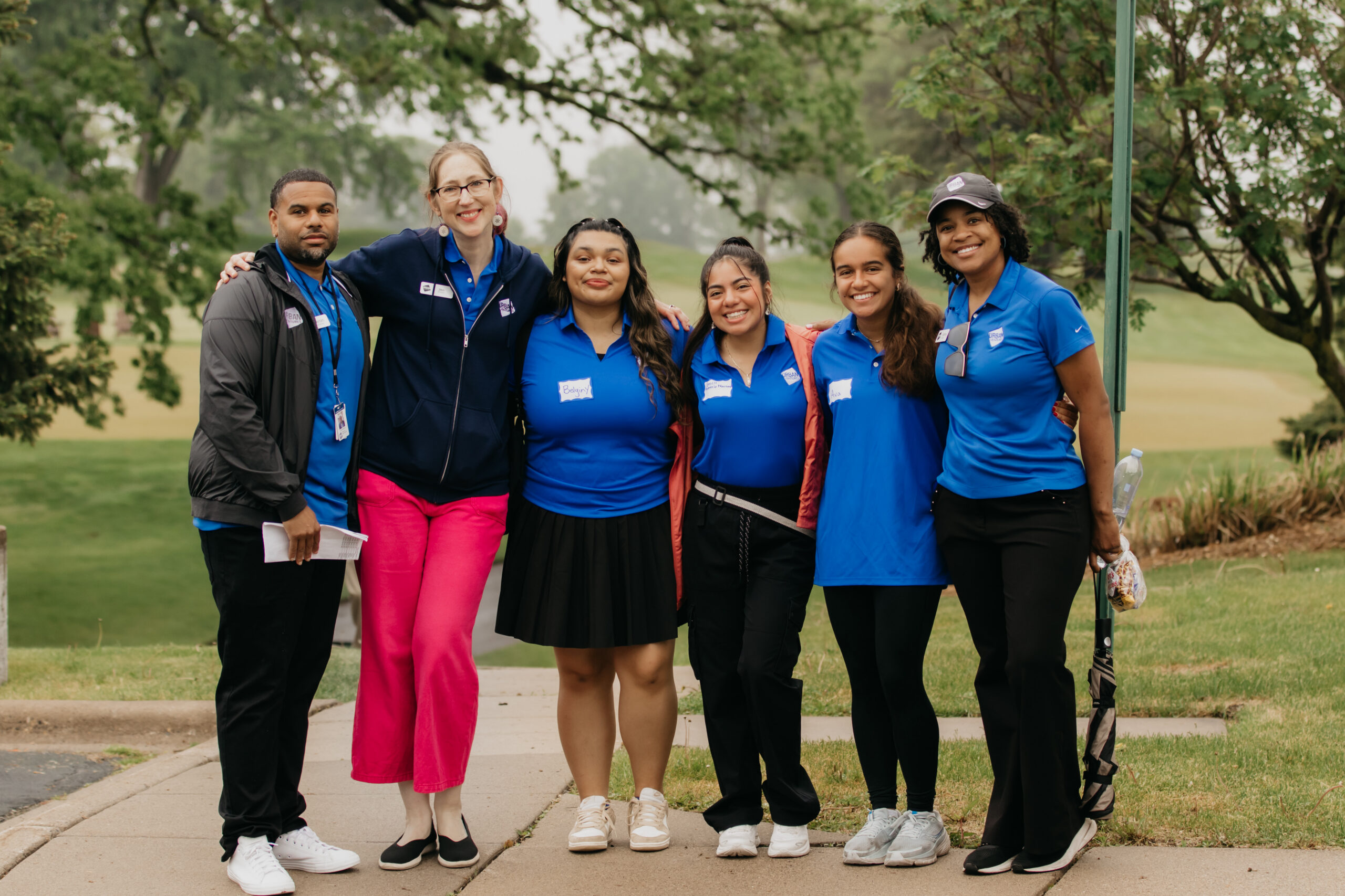Six Urban Ventures staff and volunteers in royal blue polos posing arm in arm on a paved path beside a golf course on an overcast day