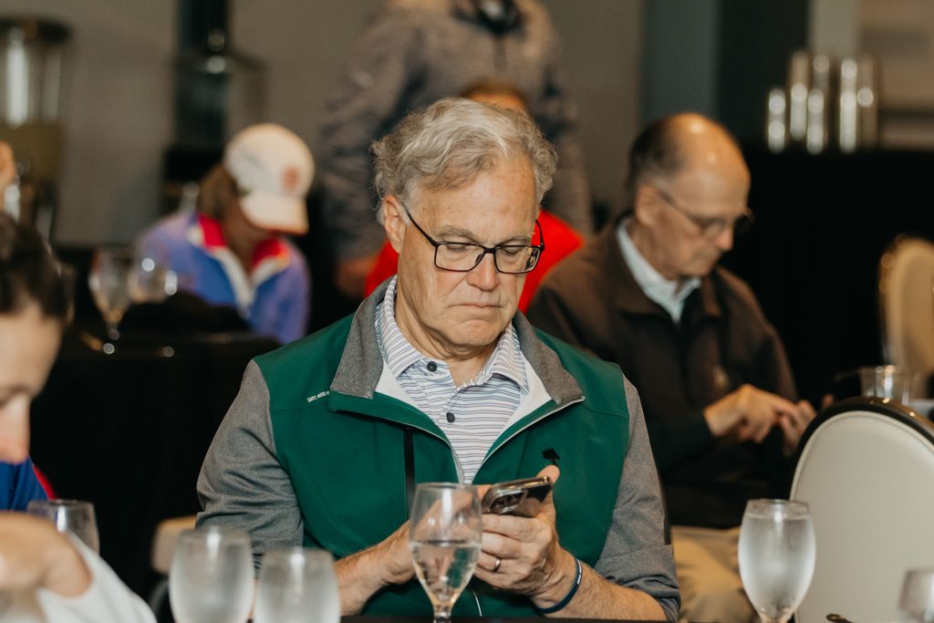 Man in glasses and a green vest checking his phone at a crowded ballroom table