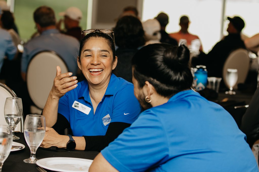 Volunteer in a blue polo laughing and gesturing at a ballroom table set with water glasses