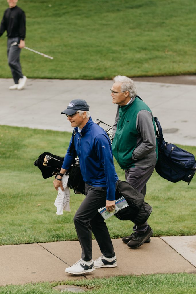 Two golfers walking a cart path with golf bags, one in a blue jacket and one in a green vest