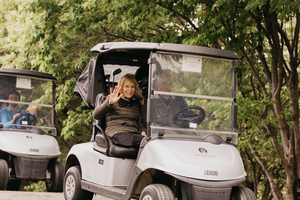 Woman in an olive jacket waving from the passenger seat of a golf cart