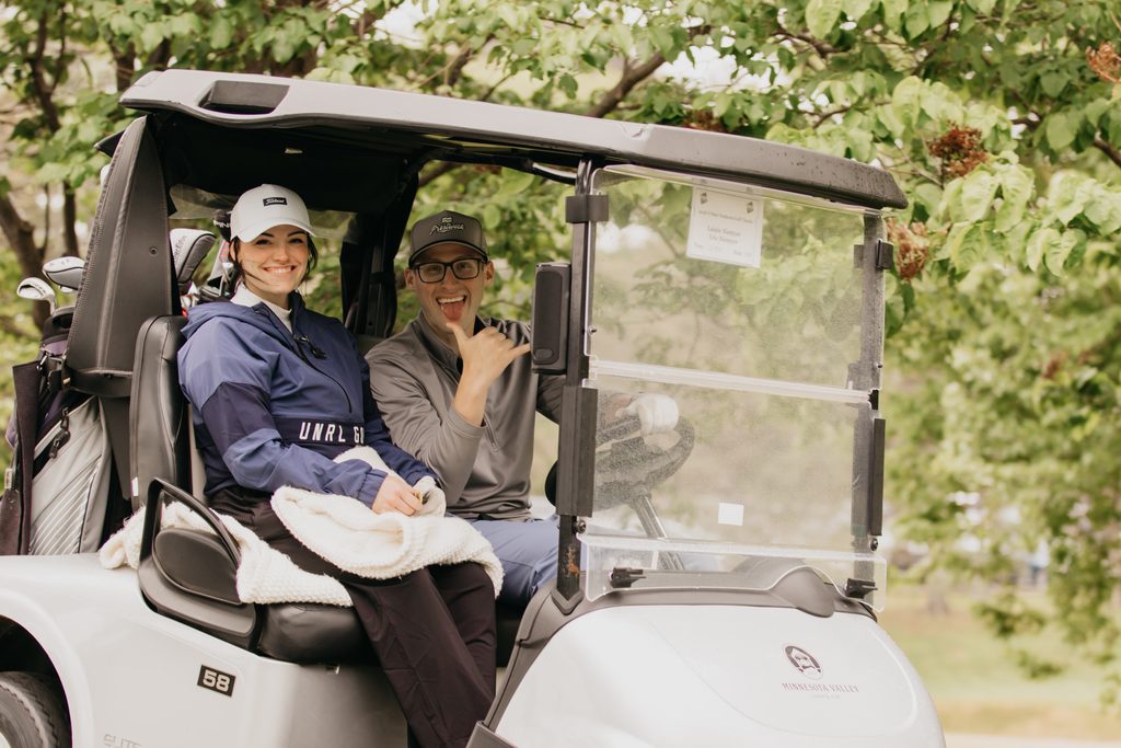 Two women in a golf cart, one in a navy jacket and white cap, smiling at the camera