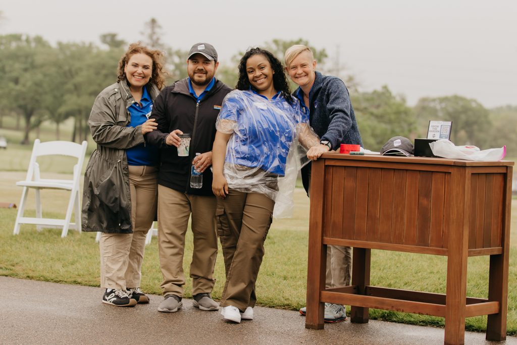 Five staff and volunteers pose beside a wooden table on a wet golf course under a gray sky