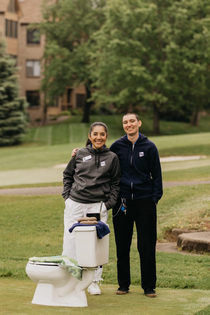 Two smiling volunteers in navy jackets standing beside a toilet prop on the grass at a golf hole