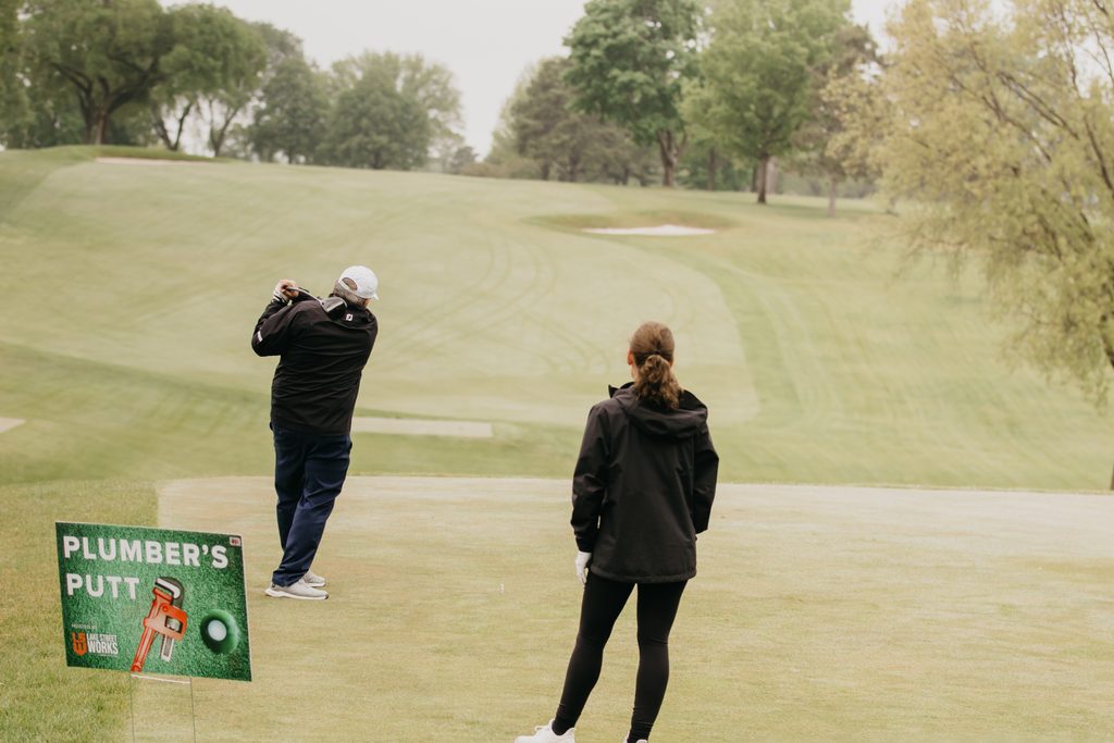 Golfer mid-swing near a Plumber’s Putt hole sign while a volunteer in a dark jacket watches