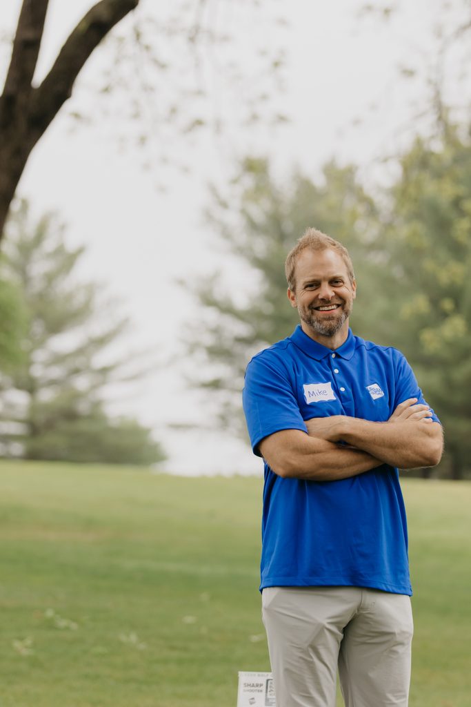 Smiling volunteer in a blue polo standing with arms crossed on the golf course