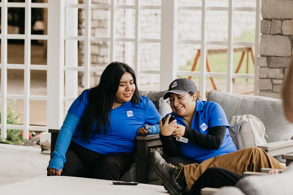 Two volunteers in blue polos laughing on an outdoor sofa while one looks at a phone