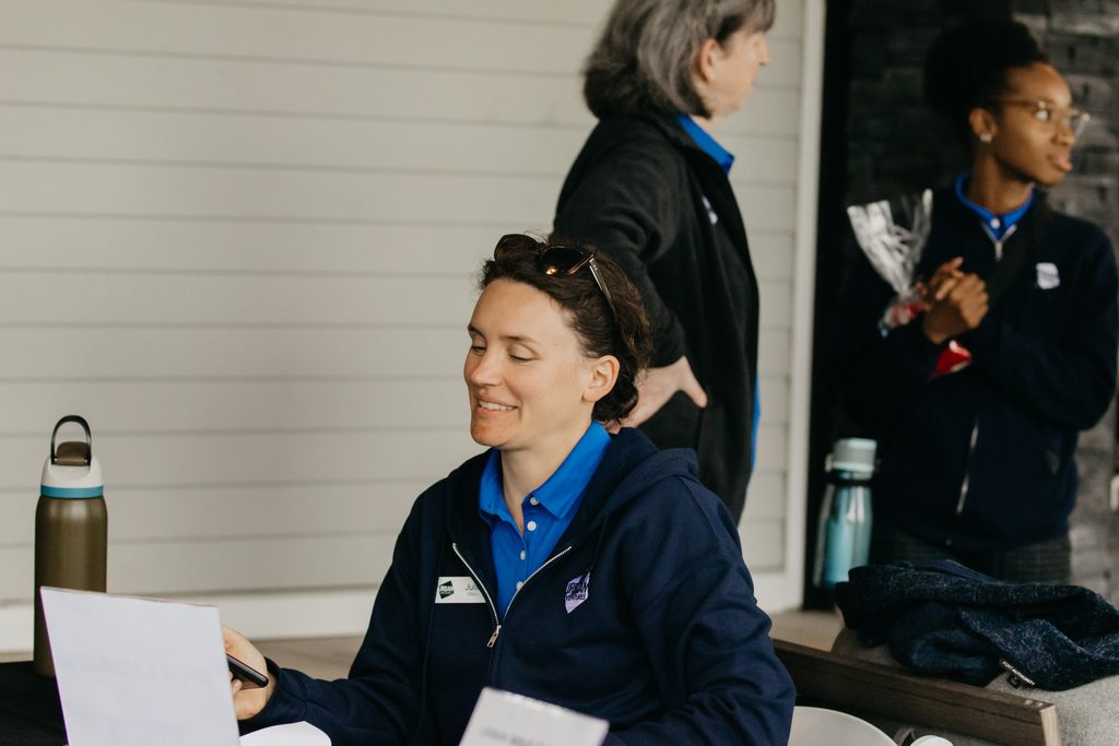 Volunteer in a navy jacket smiling as she writes at a check-in table, teammates behind her