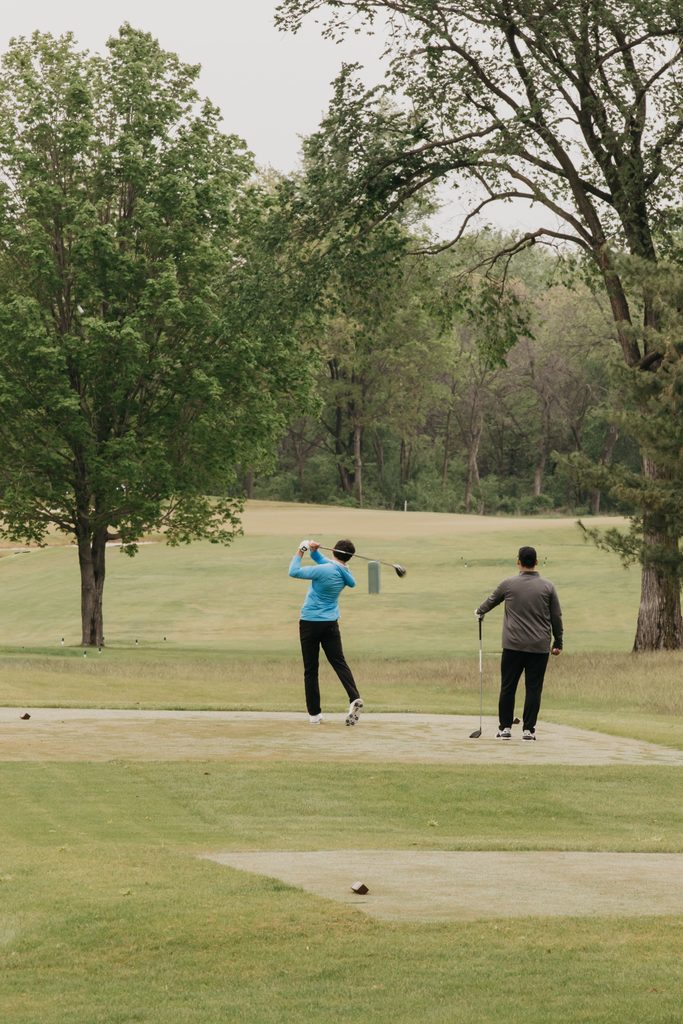 Two golfers on adjacent tee boxes, one mid-swing, with trees behind