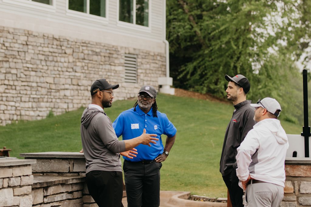 Small group of men chatting on a stone patio, one in a blue polo and the others in caps