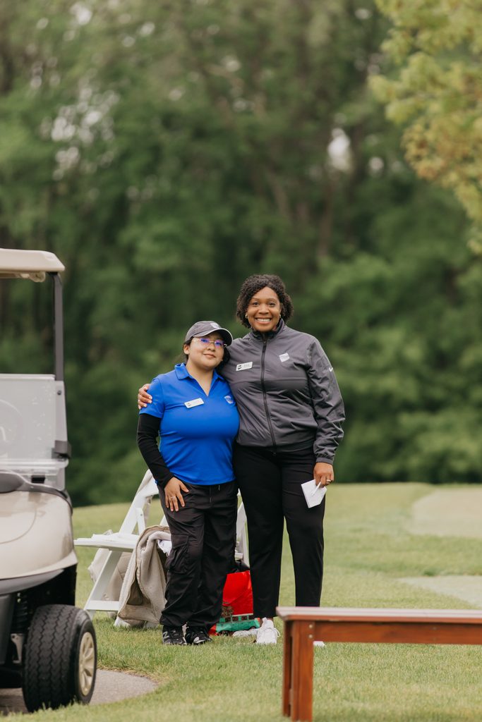 Two women posing arm in arm beside a golf cart, one in a blue polo and one in a gray jacket