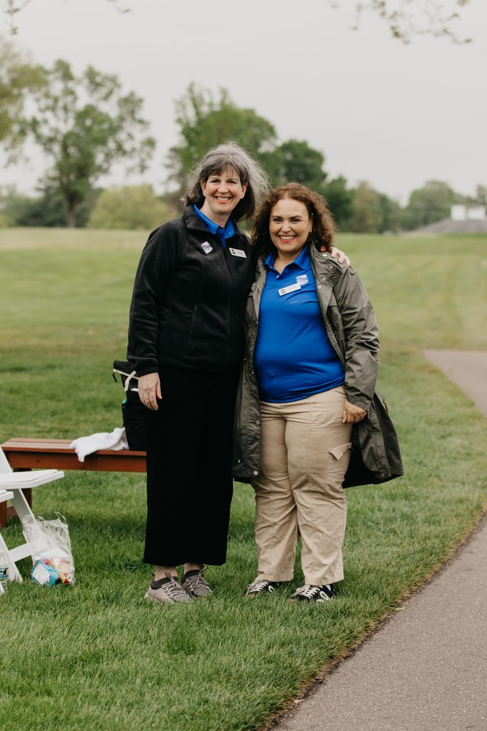 Two women smile together on a grassy path, one in a black jacket and one in a blue polo with a name tag