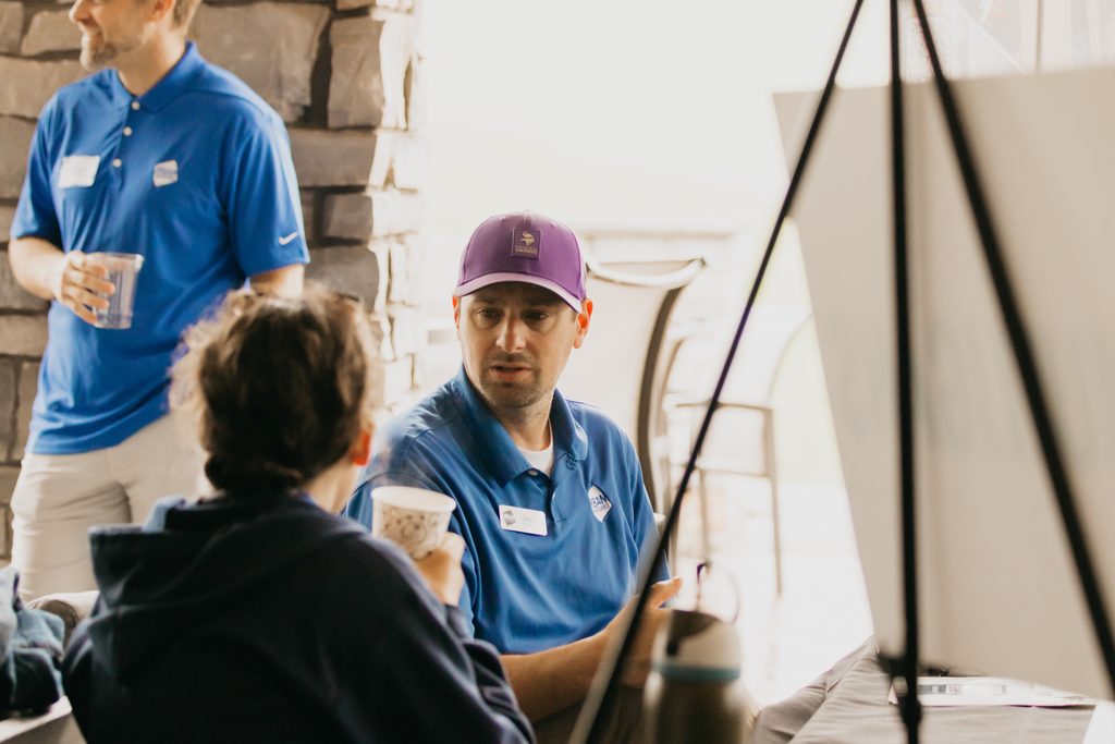 Two men seated at a patio table holding paper cups, one in a purple cap and blue polo