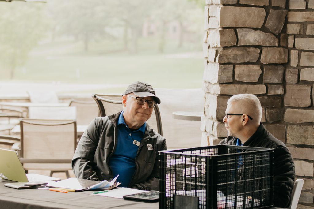Two men seated at a patio table with a laptop and papers beside a stone pillar