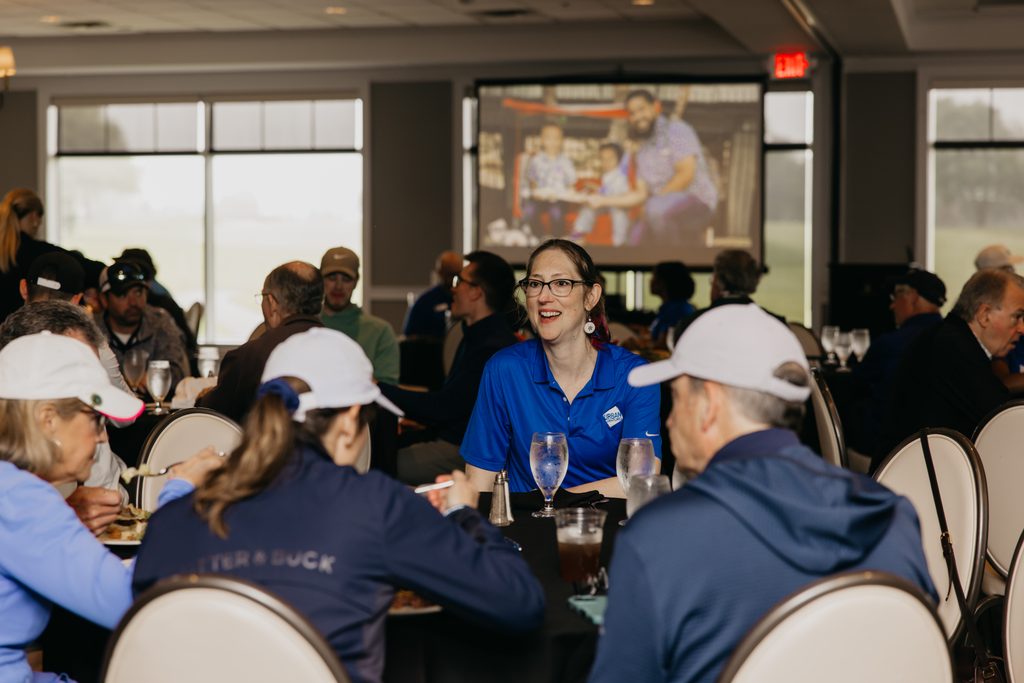 Guests at ballroom tables with a smiling volunteer in a blue polo and a slide on the screen behind