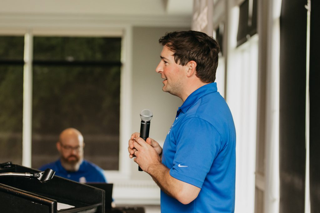Man in a blue polo speaking into a handheld microphone in a bright ballroom