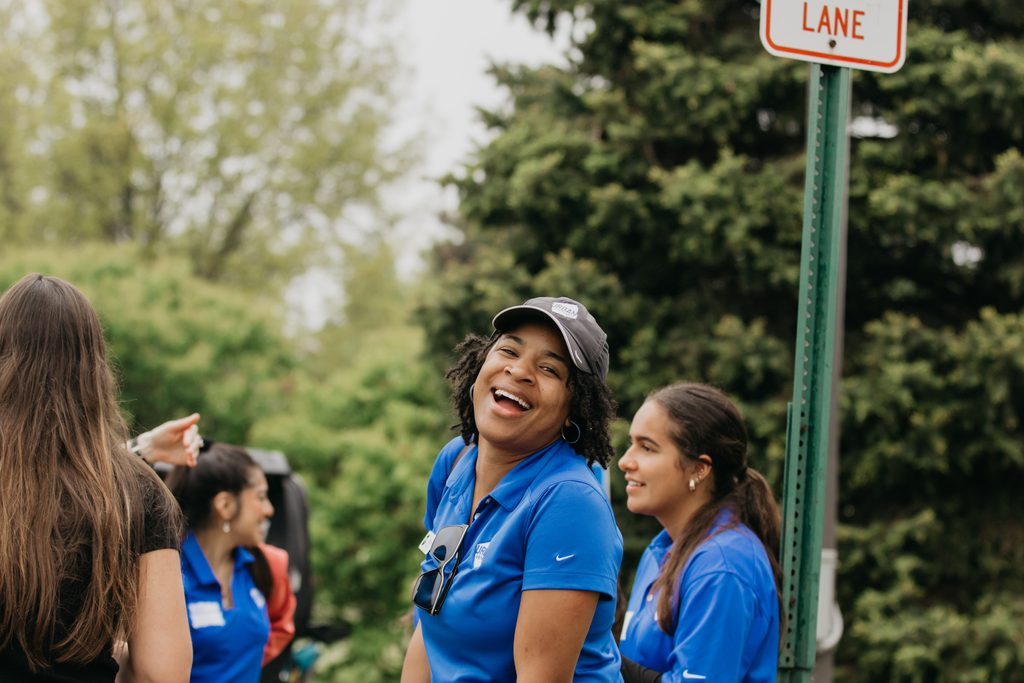 Three Urban Ventures volunteers in blue polos laughing together beside a street sign at the golf event