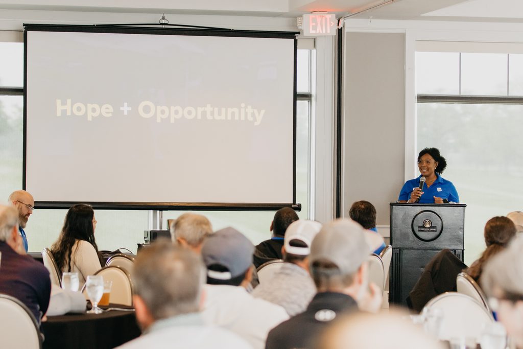 Speaker in a blue polo at a podium beside a screen reading “Hope + Opportunity” as guests watch