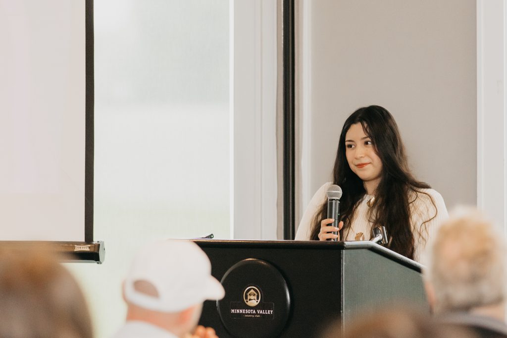 Woman with long dark hair speaking into a microphone at a podium while guests in caps listen