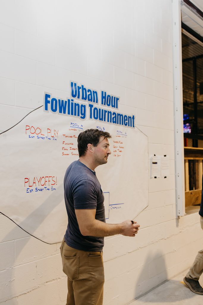 Man in a gray t-shirt stands at a white wall in front of a hand-lettered Urban Hour Fowling Tournament bracket